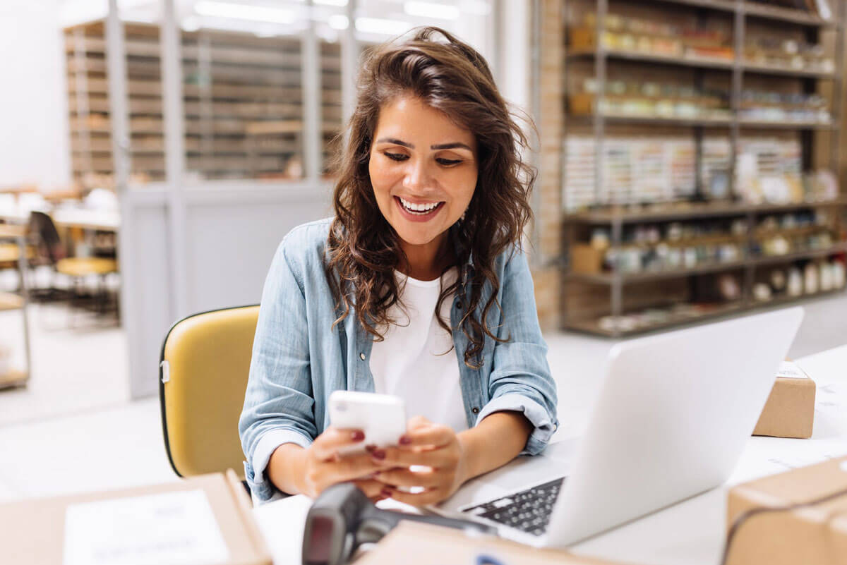 A woman smiling while using a smartphone at a workspace with a laptop