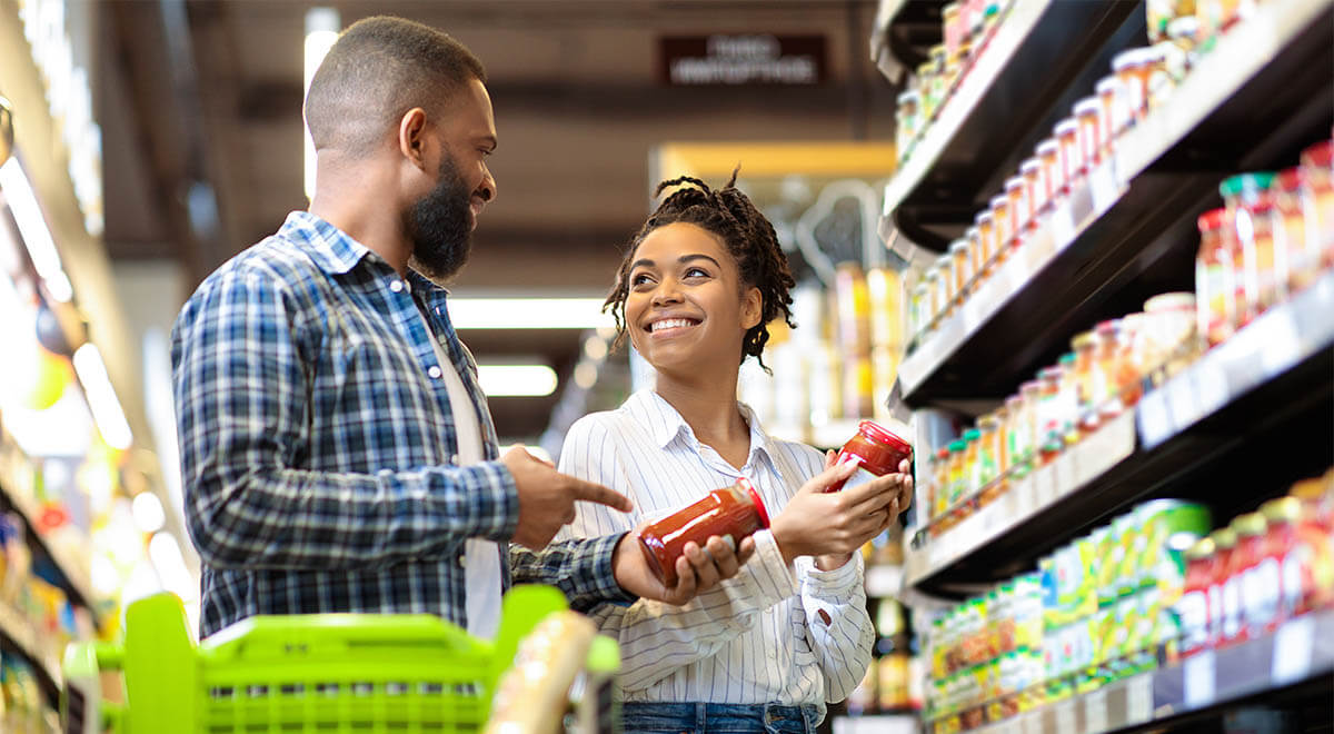 A man and woman examine food items together in a grocery store aisle, discussing their choices.