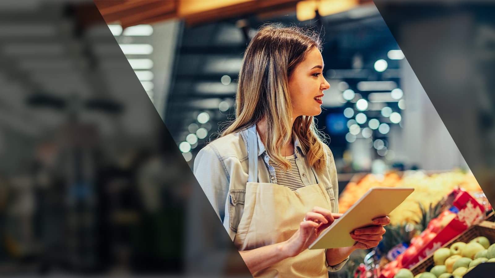 A woman stands in front of a produce section, holding a tablet and looking at the fresh fruits and vegetables.
